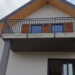 Szara, metalowa balustrada balkonowa z pionowymi szczebelkami, zamontowana na betonowej płycie balkonu w nowoczesnym domu z drewnianym podbitką dachową.