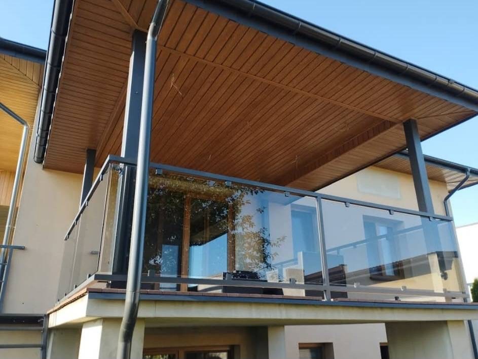 Nowoczesna balustrada szklana z grafitową ramą na balkonie domu. Widoczne odbicie drzewa w szkle, brązowe podbitka dachowa i rynna. Jasne, słoneczne otoczenie.