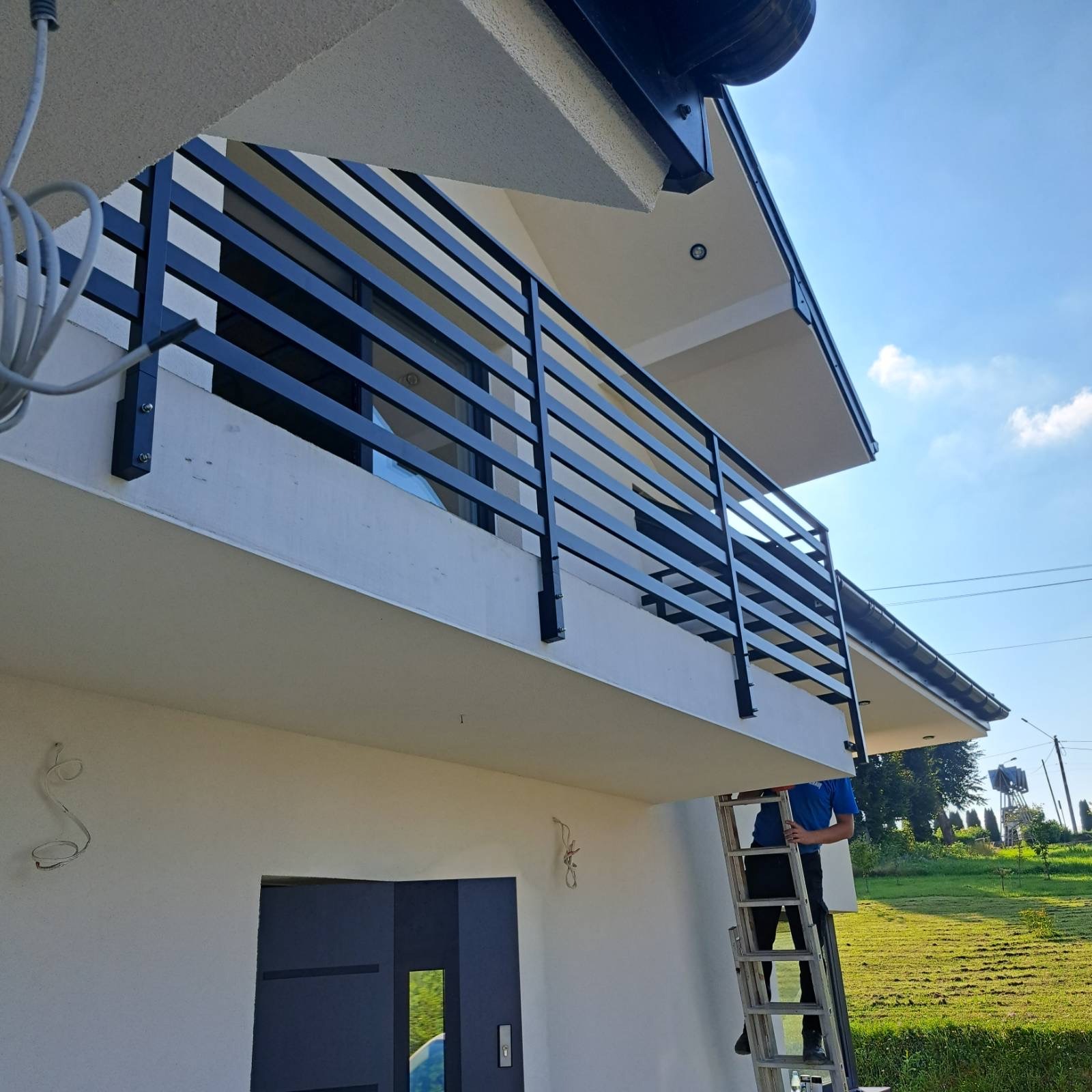 Czarna, pozioma balustrada balkonowa z metalu, zamontowana na jasnym budynku. Widok z dołu, na pierwszym planie drabina i osoba w trakcie montażu.
