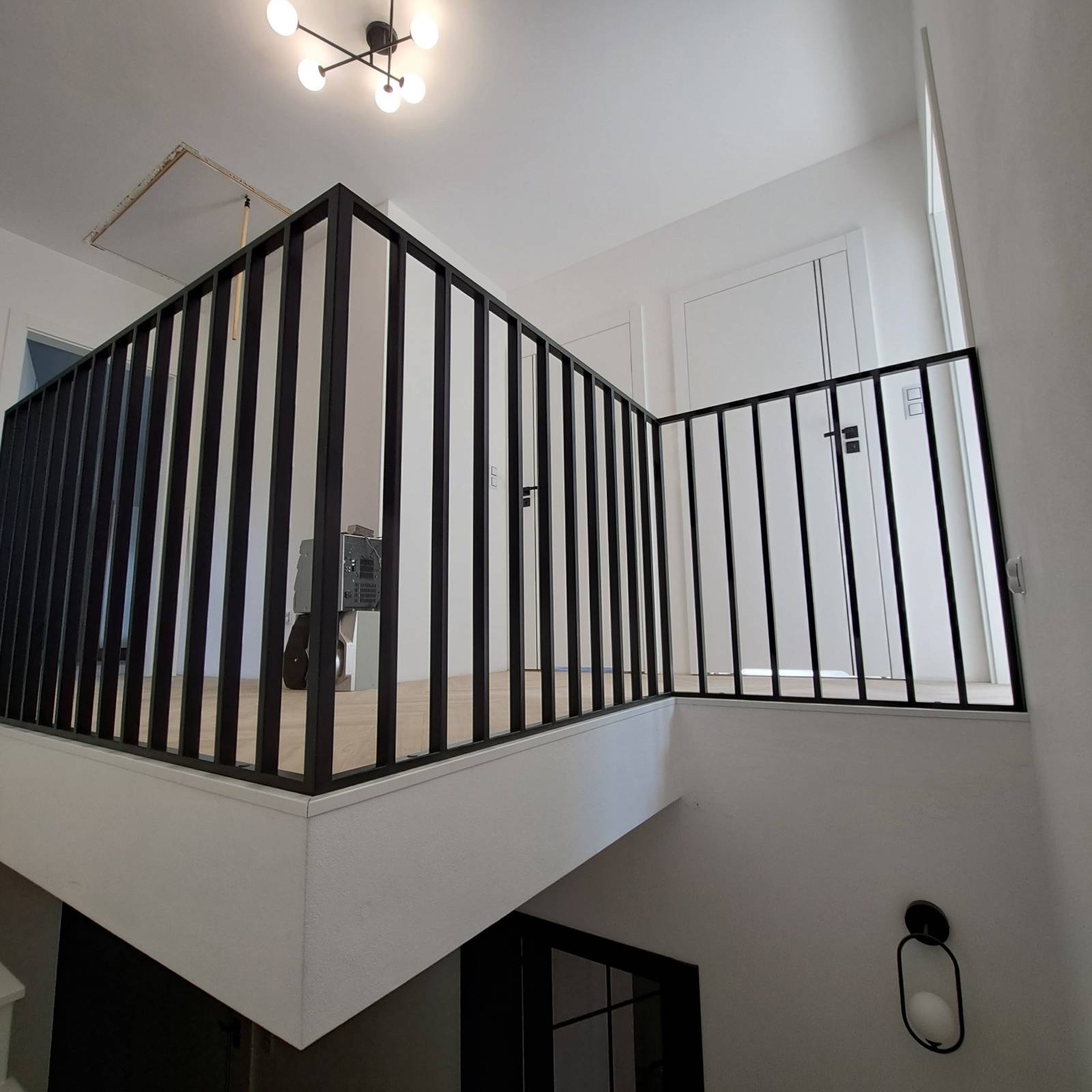 Nowoczesna, czarna balustrada metalowa z pionowymi szczeblami, zamontowana na antresoli w jasnym wnętrzu. Widoczne drzwi i lampa sufitowa.