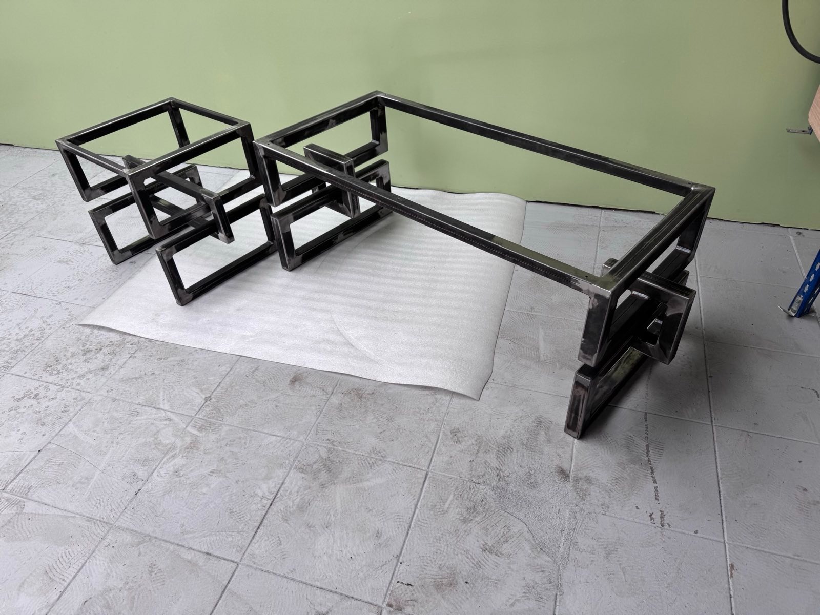 Surowa metalowa konstrukcja stolika kawowego z geometrycznym wzorem, złożona z kwadratowych profili, na białej piance. Widoczne ślady spawania i szlifowania.