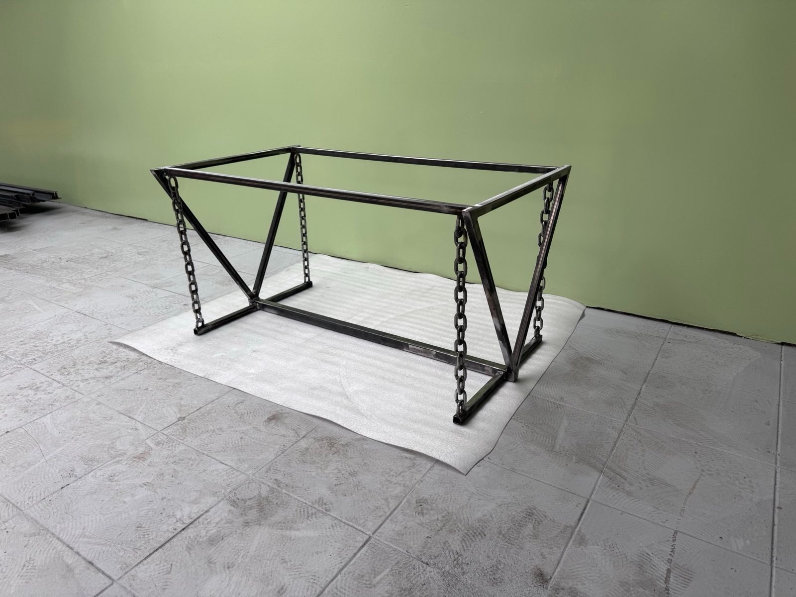 Metalowa konstrukcja stołu z łańcuchami, spawana, surowa, na białej piance. Nowoczesny design, zielona ściana w tle. Elementy spawane pod kątem.