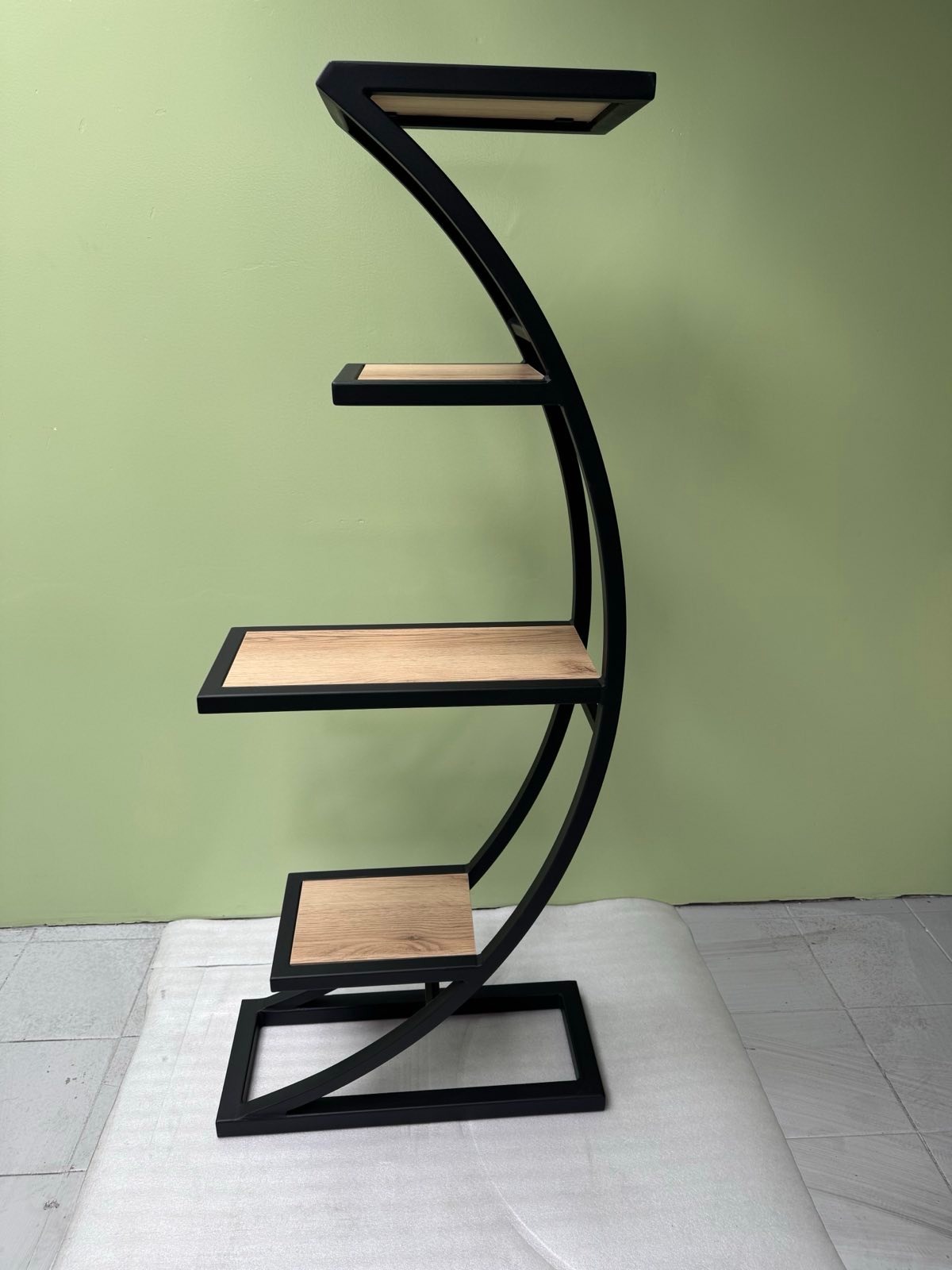 Metalowy regał o unikalnym, zakrzywionym kształcie z drewnianymi półkami na tle zielonej ściany. Czarna, matowa rama. Nowoczesny design.