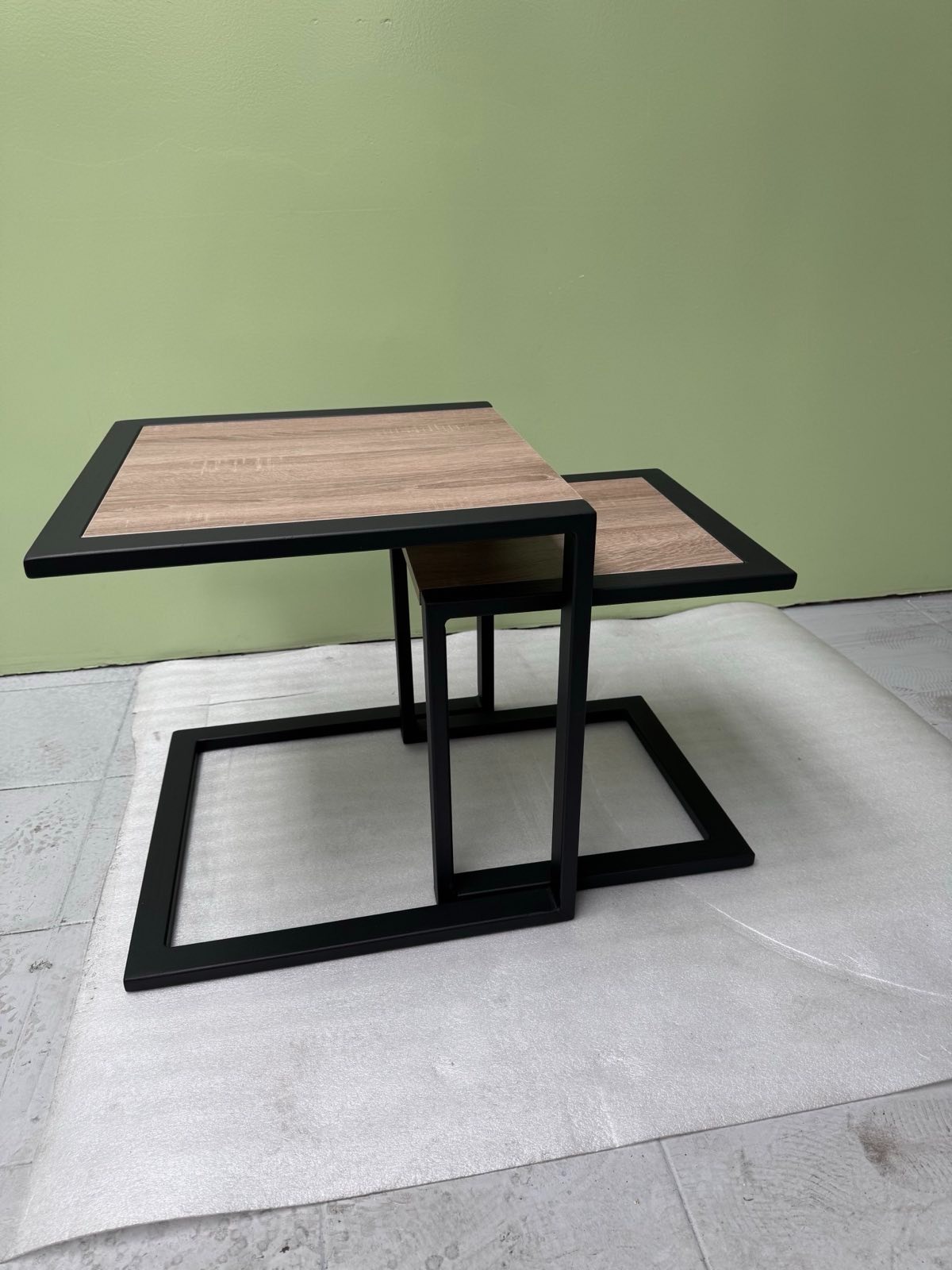 Dwa stoliki kawowe z drewnianymi blatami i czarnymi metalowymi ramami, ustawione jeden obok drugiego. Minimalistyczny design na tle zielonej ściany.