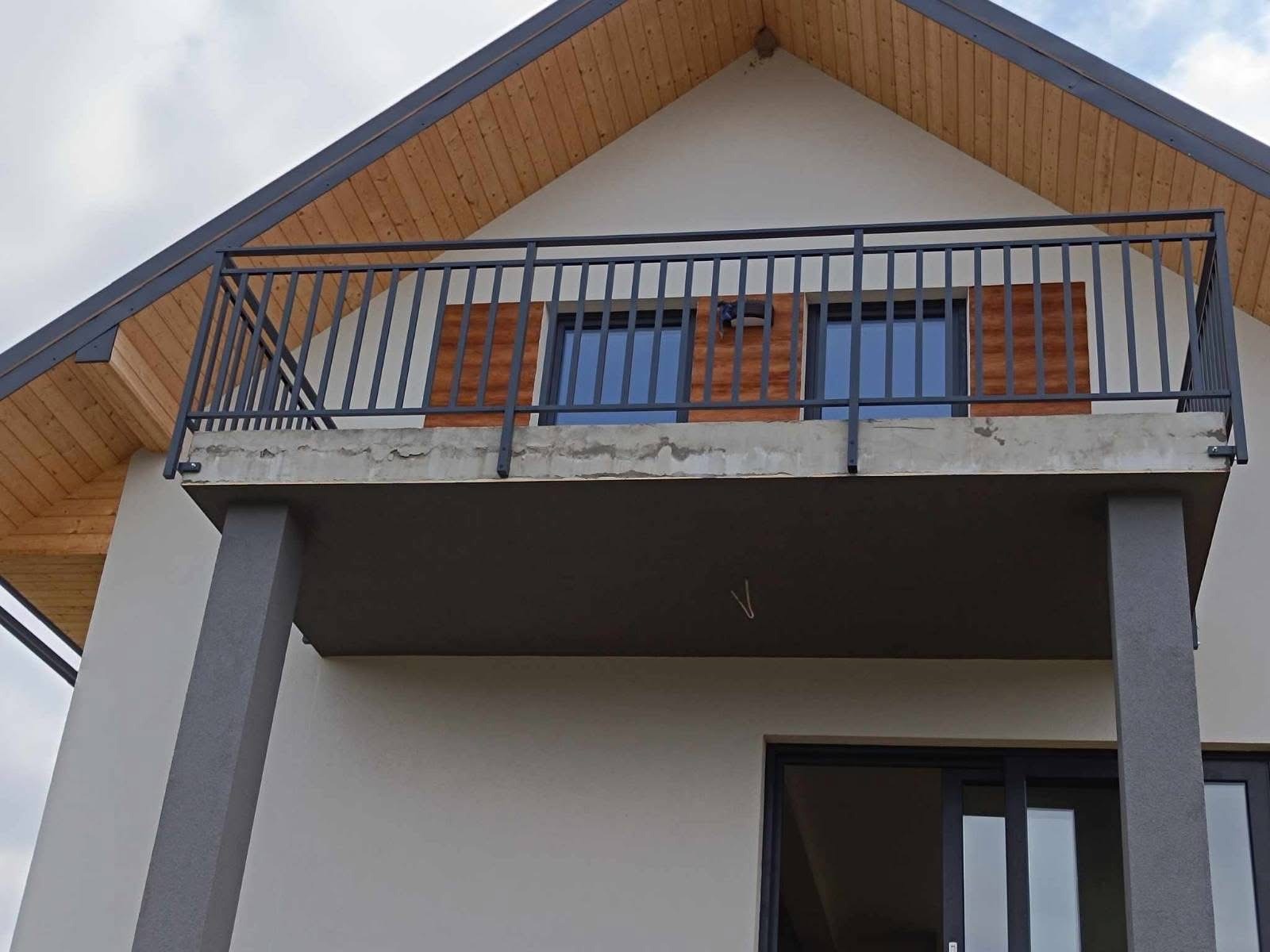 Szara, metalowa balustrada balkonowa z pionowymi szczebelkami, zamontowana na betonowej płycie balkonu w nowoczesnym domu z drewnianym podbitką dachową.