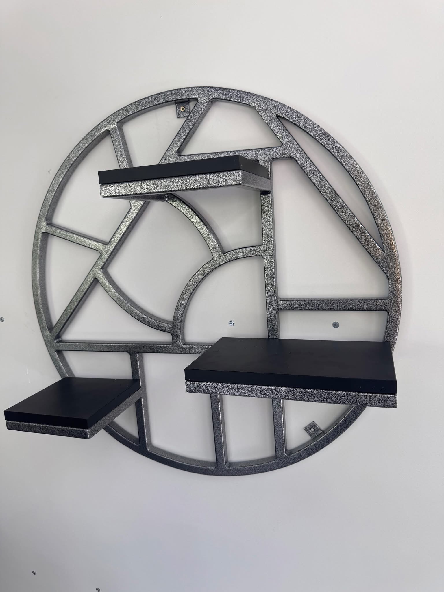 Metalowa półka ścienna w kształcie okręgu z geometrycznym wzorem i trzema czarnymi półkami, zamontowana na białej ścianie. Wykończenie w kolorze srebrnym.