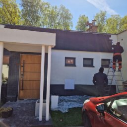 Ludovic Stoica - Malowanie elewacji domu jednorodzinnego: dwóch pracowników na drabinie, jeden maluje ścianę, drugi obserwuje. Czerwony samochód na pierwszym planie.