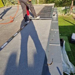 Ludovic Stoica - Prace dekarskie na płaskim dachu: dekarz w kombinezonie układa papę, widoczna listwa z logo IZBUD i fragment rynny, zieleń w tle.