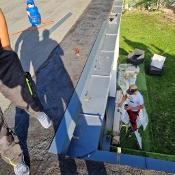 Ludovic Stoica - Prace na dachu: malowanie obróbek blacharskich przy użyciu drabiny, widoczny gazowy palnik dekarski, narzędzia i fragment trawnika z meblami ogrodowymi.