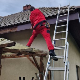 Ludovic Stoica - Pracownik w czerwonym kombinezonie na drabinie, montuje obróbkę blacharską dachu. Widoczny fragment dachu z dachówką ceramiczną i fragment ściany budynku.