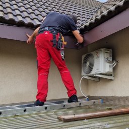 Ludovic Stoica - Pracownik w czerwonych spodniach montuje brązowe obróbki blacharskie przy dachu z dachówki ceramicznej, stojąc na metalowej drabinie. Widoczny klimatyzator.