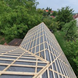 Ludovic Stoica - Konstrukcja dachu z łatami i membraną w trakcie budowy, widok z góry na tle zieleni. Częściowo widoczna sylwetka osoby pracującej na dachu.