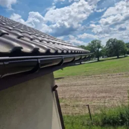 Brązowe rynny zamontowane na dachu z dachówki ceramicznej, widok z dołu na tle błękitnego nieba i zielonego krajobrazu. Widoczny fragment elewacji budynku.