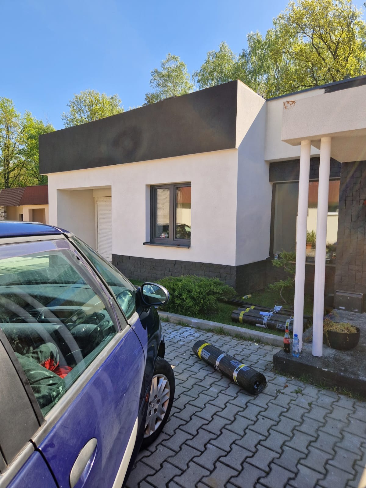 Nowoczesny dom jednorodzinny z czarnym dachem i białymi ścianami. Na pierwszym planie zwinięte rolki papy dachowej obok zaparkowanego, niebieskiego auta.