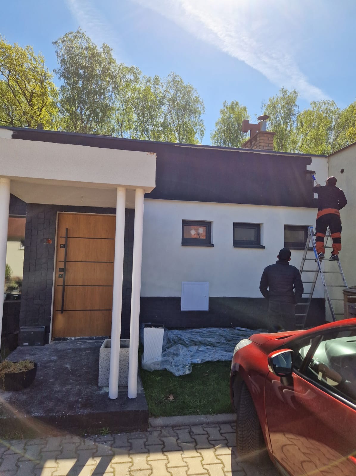 Malowanie elewacji domu jednorodzinnego: dwóch pracowników na drabinie, jeden maluje ścianę, drugi obserwuje. Czerwony samochód na pierwszym planie.