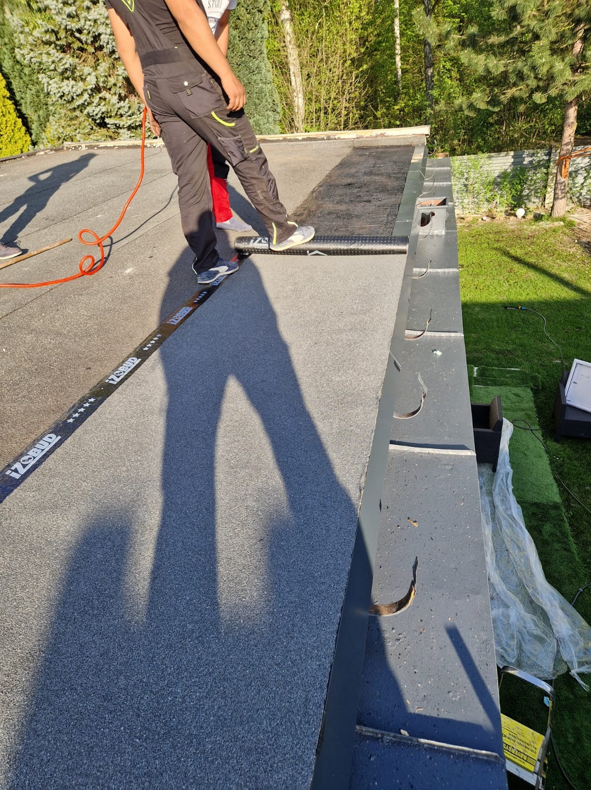 Prace dekarskie na płaskim dachu: dekarz w kombinezonie układa papę, widoczna listwa z logo IZBUD i fragment rynny, zieleń w tle.