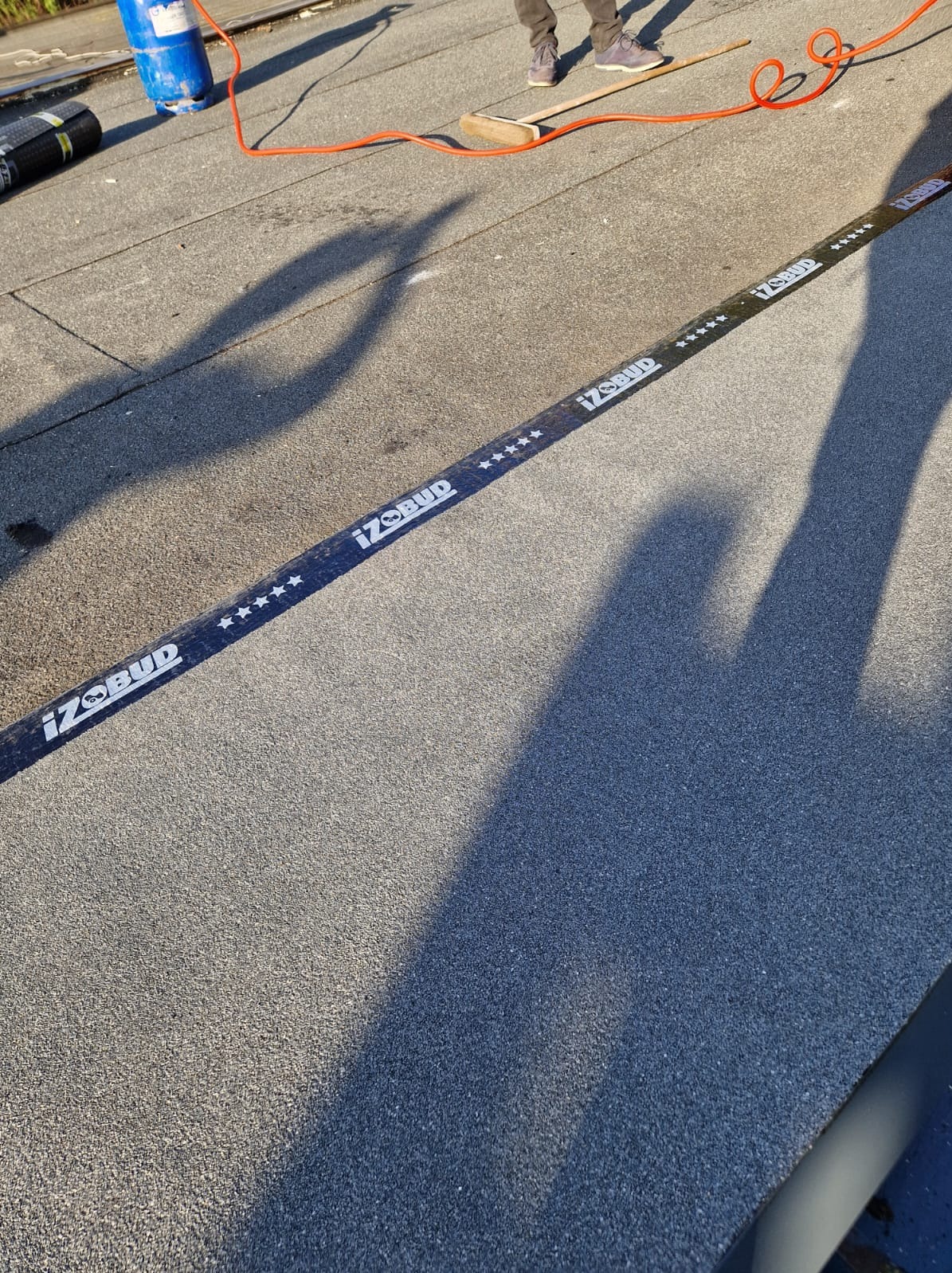 Praca na dachu: widoczny fragment dachu z papy termozgrzewalnej z logo firmy, butla gazowa, palnik i cień osoby podczas wykonywania prac dekarskich.