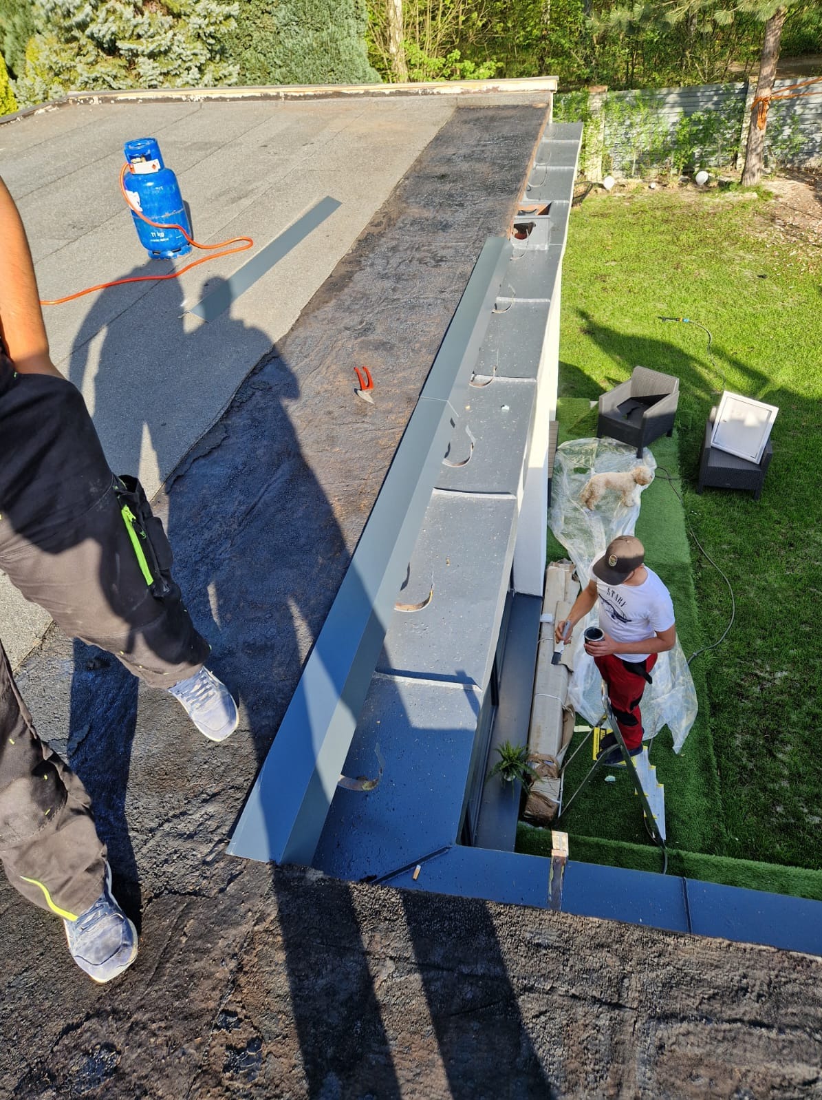 Prace na dachu: malowanie obróbek blacharskich przy użyciu drabiny, widoczny gazowy palnik dekarski, narzędzia i fragment trawnika z meblami ogrodowymi.