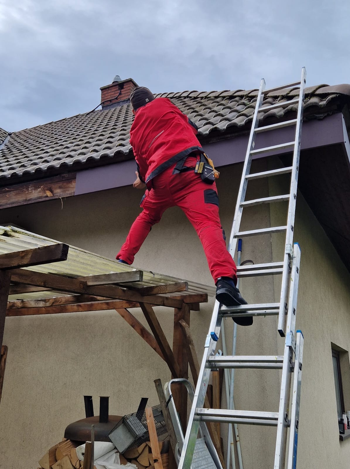 Pracownik w czerwonym kombinezonie na drabinie, montuje obróbkę blacharską dachu. Widoczny fragment dachu z dachówką ceramiczną i fragment ściany budynku.