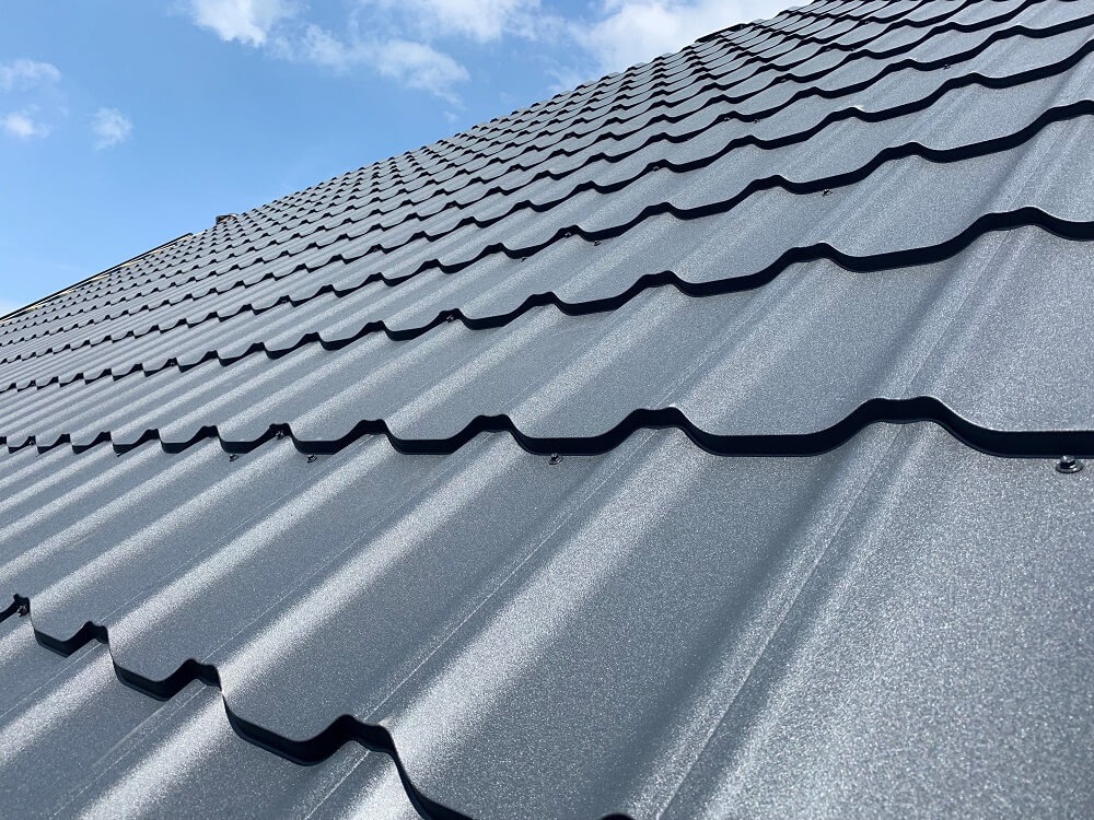 Nowoczesny, grafitowy dach z blachy trapezowej, widoczny pod kątem. Na tle błękitne niebo z delikatnymi obłokami.  Widoczne mocowania.