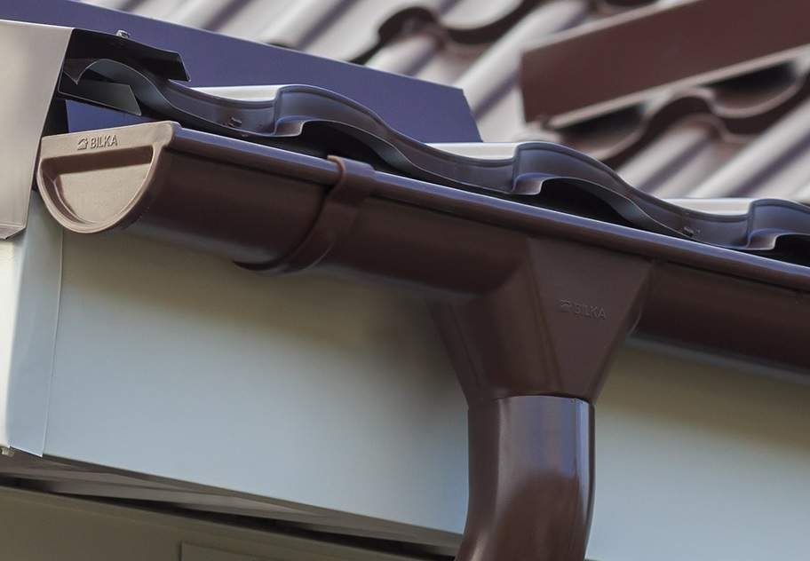 Czekoladowa rynna z logo Bilka, zamontowana przy dachu z dachówki ceramicznej w Katowicach. Widoczny fragment obróbki blacharskiej i orynnowania.