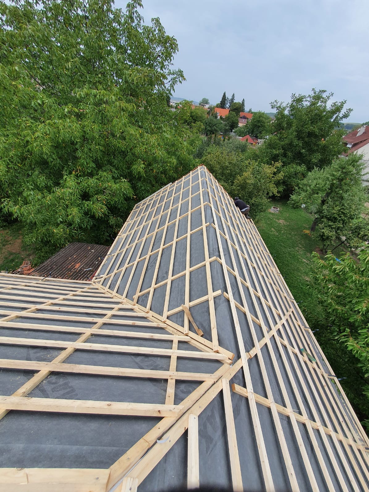 Konstrukcja dachu z łatami i membraną w trakcie budowy, widok z góry na tle zieleni. Częściowo widoczna sylwetka osoby pracującej na dachu.