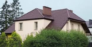 Dwupiętrowy dom z brązowym dachem krytym dachówką i beżową elewacją, otoczony zielenią. Widoczny komin i fragment dachu sąsiedniego budynku.