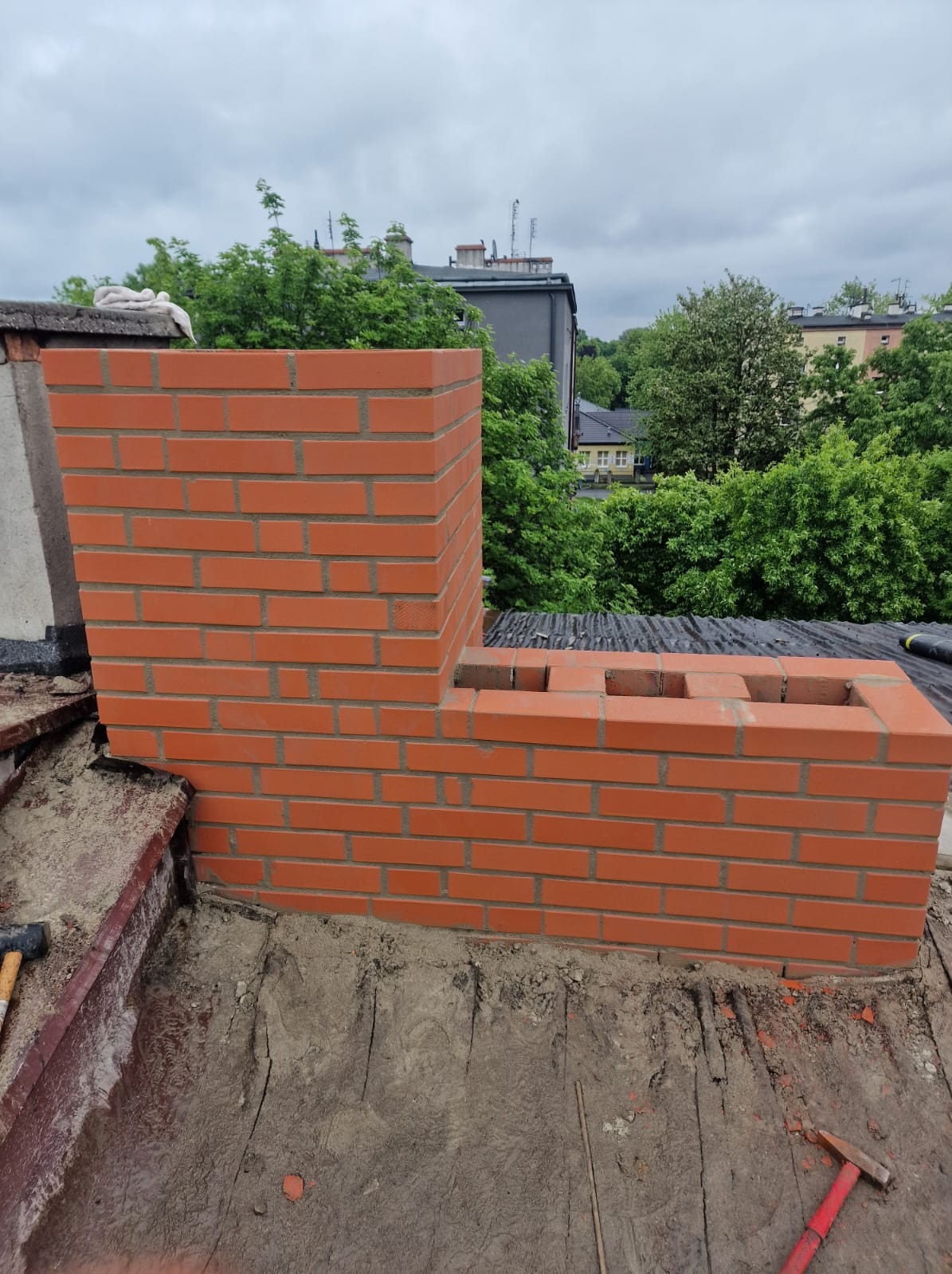 Ceglany komin w trakcie budowy na dachu, z widocznymi narzędziami i otoczeniem miejskiej zabudowy w tle. Komin z czerwonej cegły.
