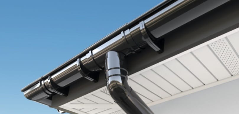 Błyszcząca, czarna rynna z odpływem na tle błękitnego nieba i białego podbitki dachowej. Nowoczesny system orynnowania, precyzyjny montaż.