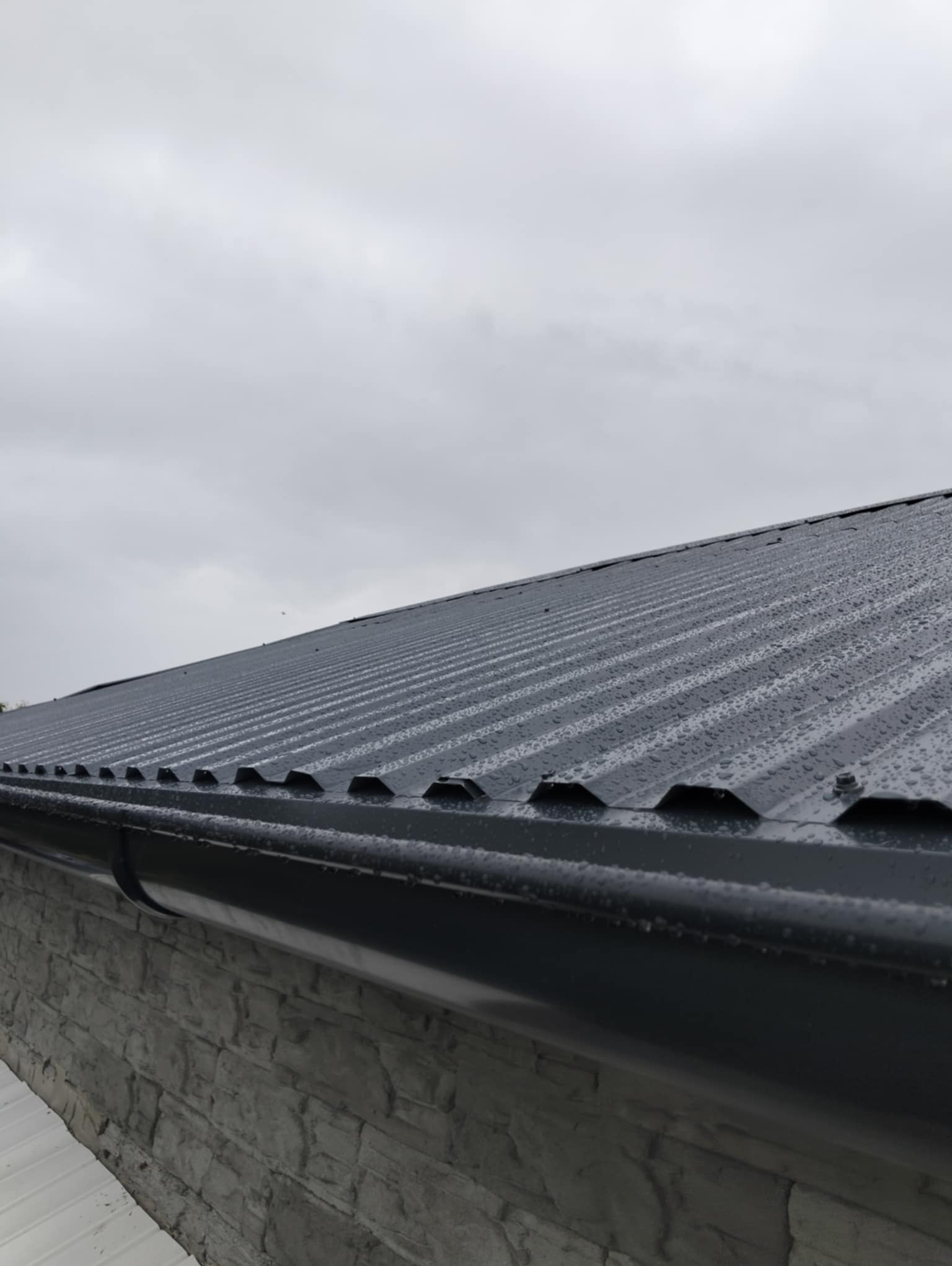 Szary, blaszany dach z nową rynną w deszczu, widok z dołu. Na pierwszym planie fragment kamiennej ściany budynku. Pochmurne niebo w tle.