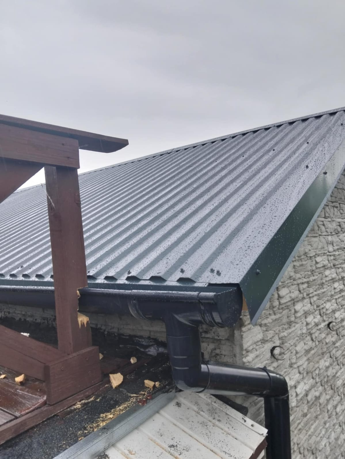 Szary dach z blachy trapezowej, z zamontowaną, czarną rynną, fragment drewnianej konstrukcji i kamiennej ściany budynku w tle, pochmurny dzień.