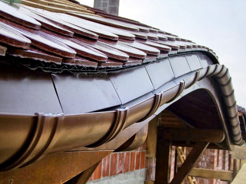 Brązowa rynna zamontowana na dachu z dachówki ceramicznej. Widoczne elementy konstrukcji drewnianej i ceglany fragment ściany budynku.