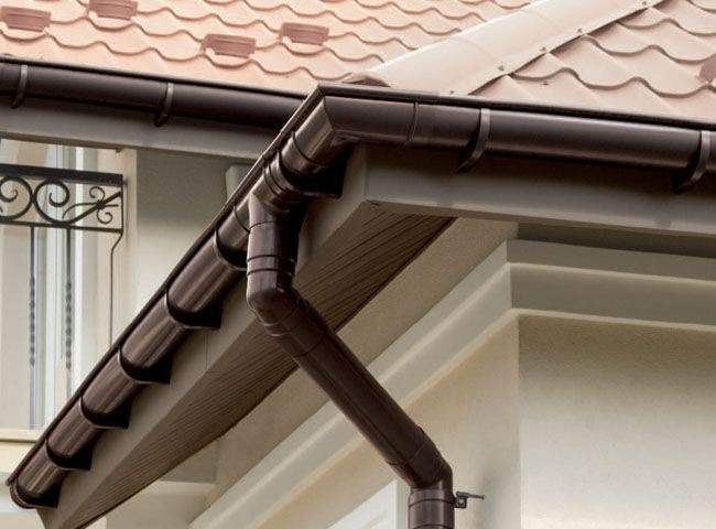 Brązowe rynny zamontowane na budynku z beżową elewacją i dachem z dachówki w odcieniach brązu. Widoczne elementy dekoracyjne okna.
