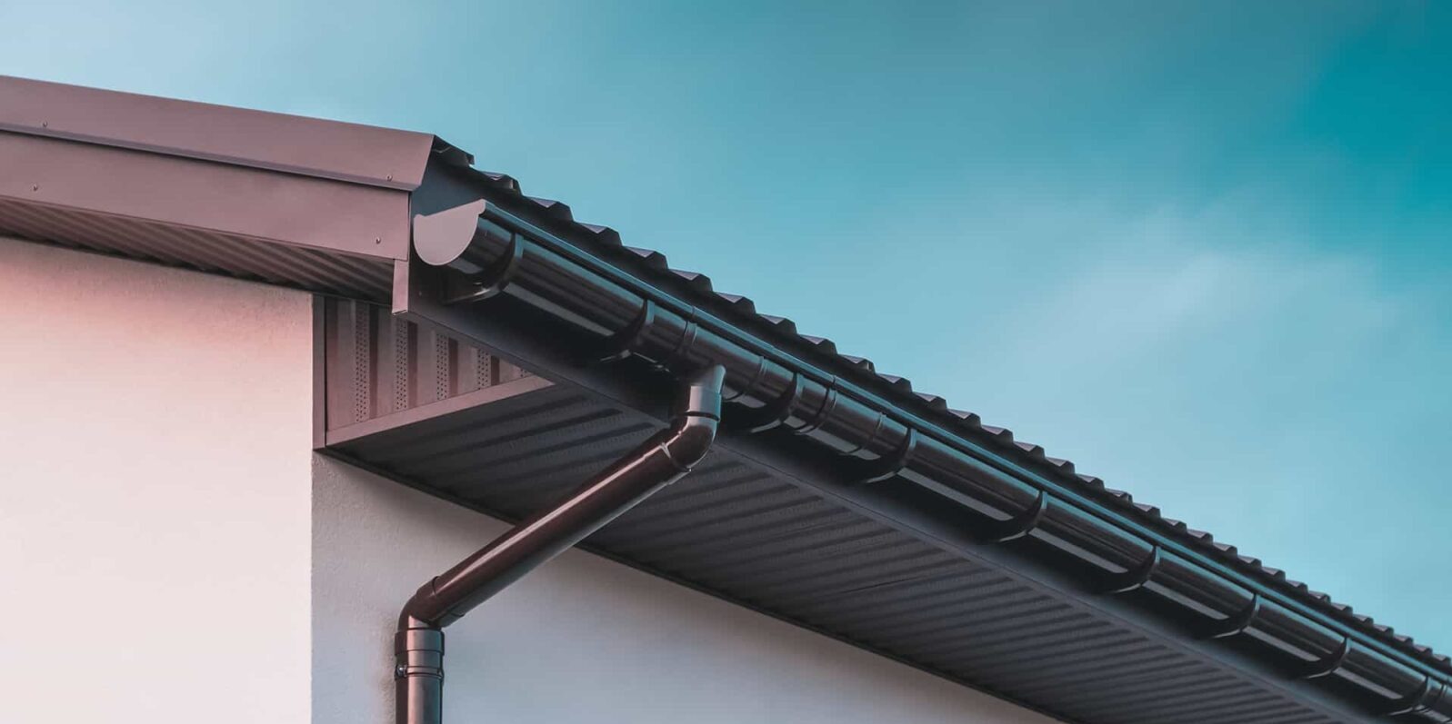 Ciemnobrązowe orynnowanie dachu domu na tle błękitnego nieba. Widoczne elementy mocujące rynny. Estetyczne wykończenie dachu.