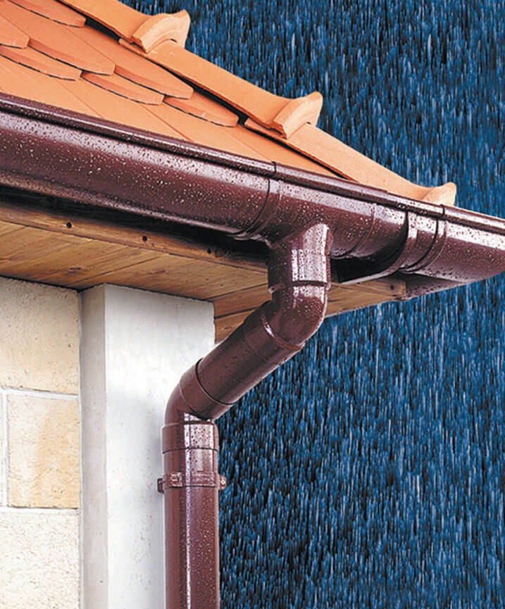 Ciemnobrązowe rynny z kroplami deszczu na tle spadającego deszczu, zamontowane przy pomarańczowym dachu z dachówki ceramicznej i białej elewacji budynku.