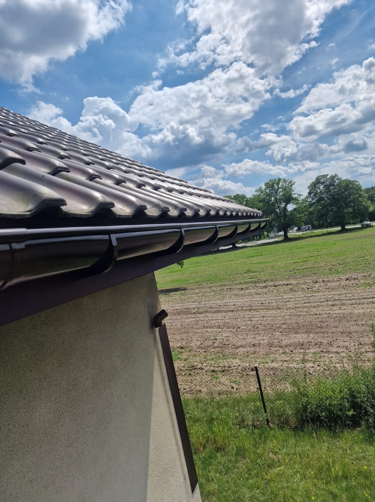 Brązowe rynny zamontowane na dachu z dachówki ceramicznej, widok z dołu na tle błękitnego nieba i zielonego krajobrazu. Widoczny fragment elewacji budynku.