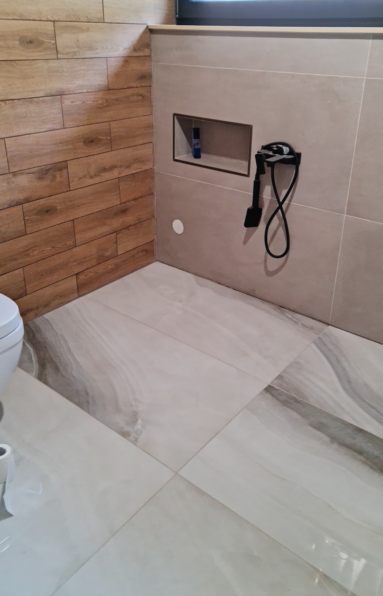 Wykończona łazienka z płytkami imitującymi drewno i marmur. W ścianie wnęka z kosmetykiem i czarny zestaw prysznicowy. Jasne, nowoczesne wnętrze.