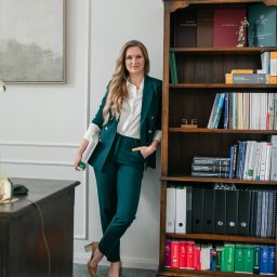 Kancelaria Adwokacka Adwokat Magdalena Chomontowska - Chełm - Adwokatka w zielonym garniturze pozuje obok regału z książkami prawniczymi w jasnym biurze. Na biurku lampa i dokumenty. Profesjonalny wizerunek prawnika.