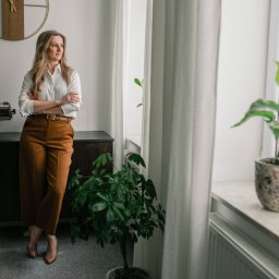 Kancelaria Adwokacka Adwokat Magdalena Chomontowska - Chełm - Portret kobiety w jasnej koszuli i brązowych spodniach, stojącej w biurze z maszyną do pisania i roślinami doniczkowymi przy oknie, spoglądającej w dal.