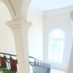 Jasne wnętrze z łukami i schodami. Balustrada z kutego żelaza, okno łukowe. Dwa krzesła z zielonymi siedziskami. Neutralna kolorystyka ścian i podłóg.