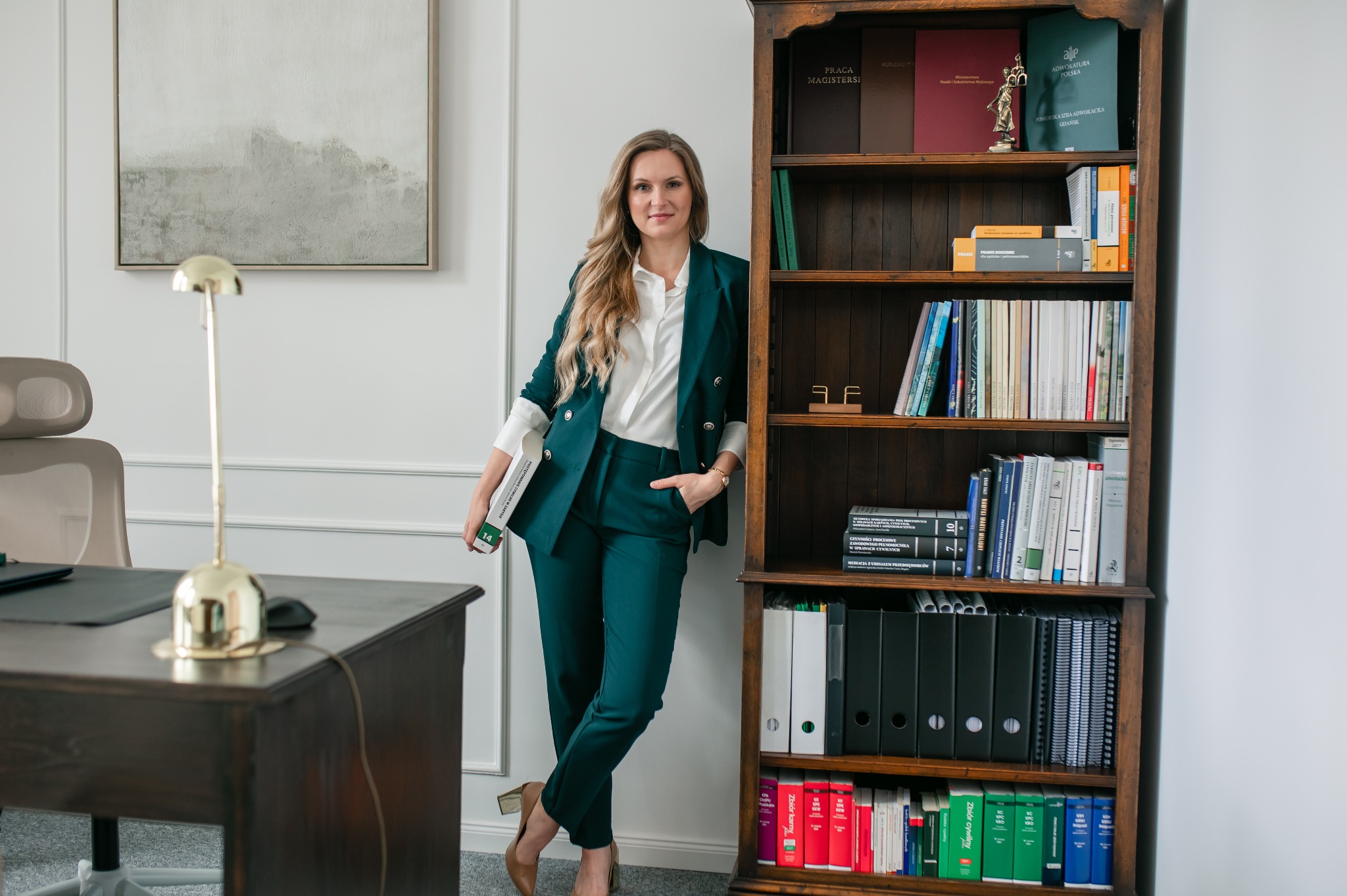 Adwokatka w zielonym garniturze pozuje obok regału z książkami prawniczymi w jasnym biurze. Na biurku lampa i dokumenty. Profesjonalny wizerunek prawnika.