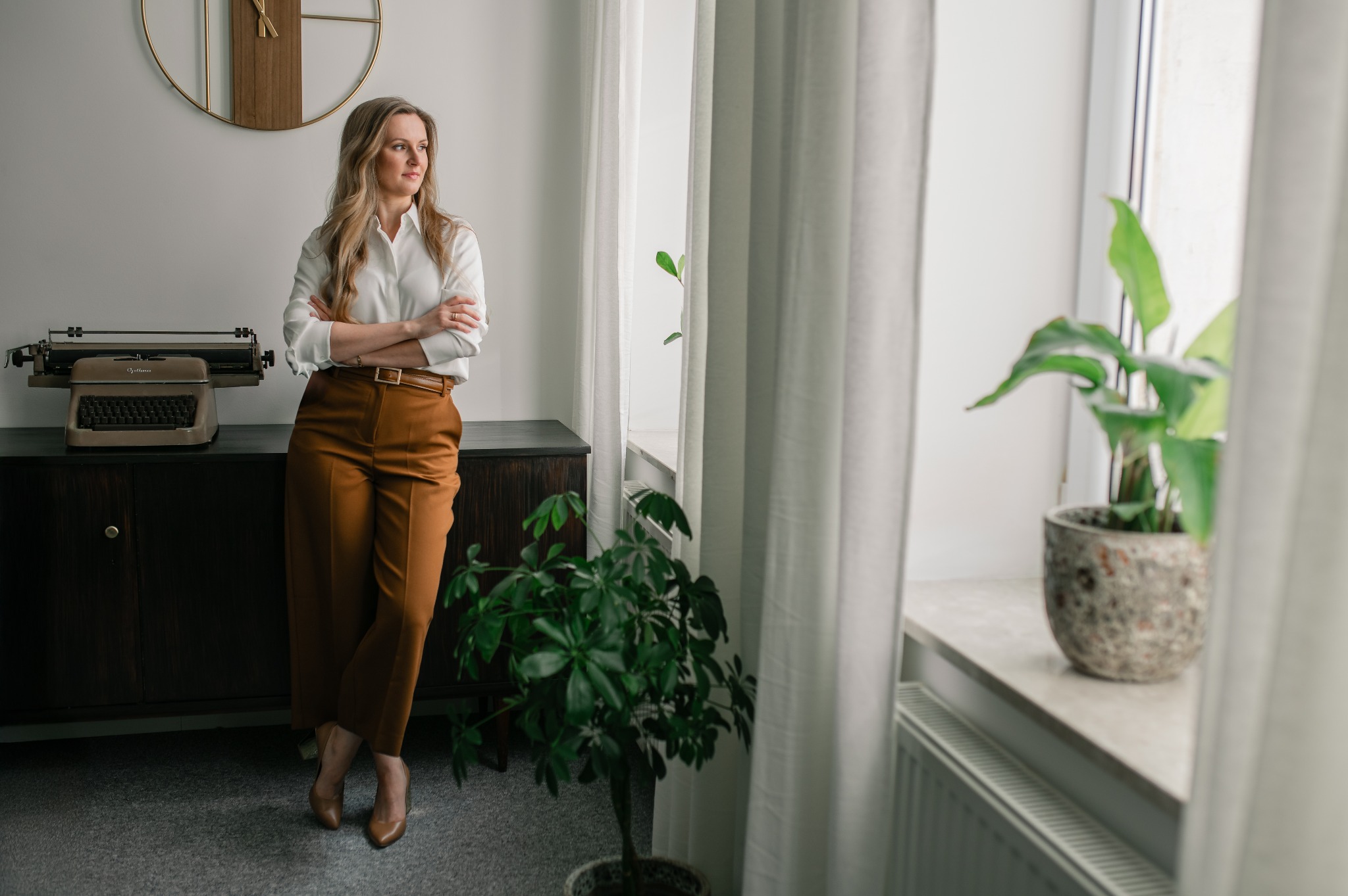 Portret kobiety w jasnej koszuli i brązowych spodniach, stojącej w biurze z maszyną do pisania i roślinami doniczkowymi przy oknie, spoglądającej w dal.
