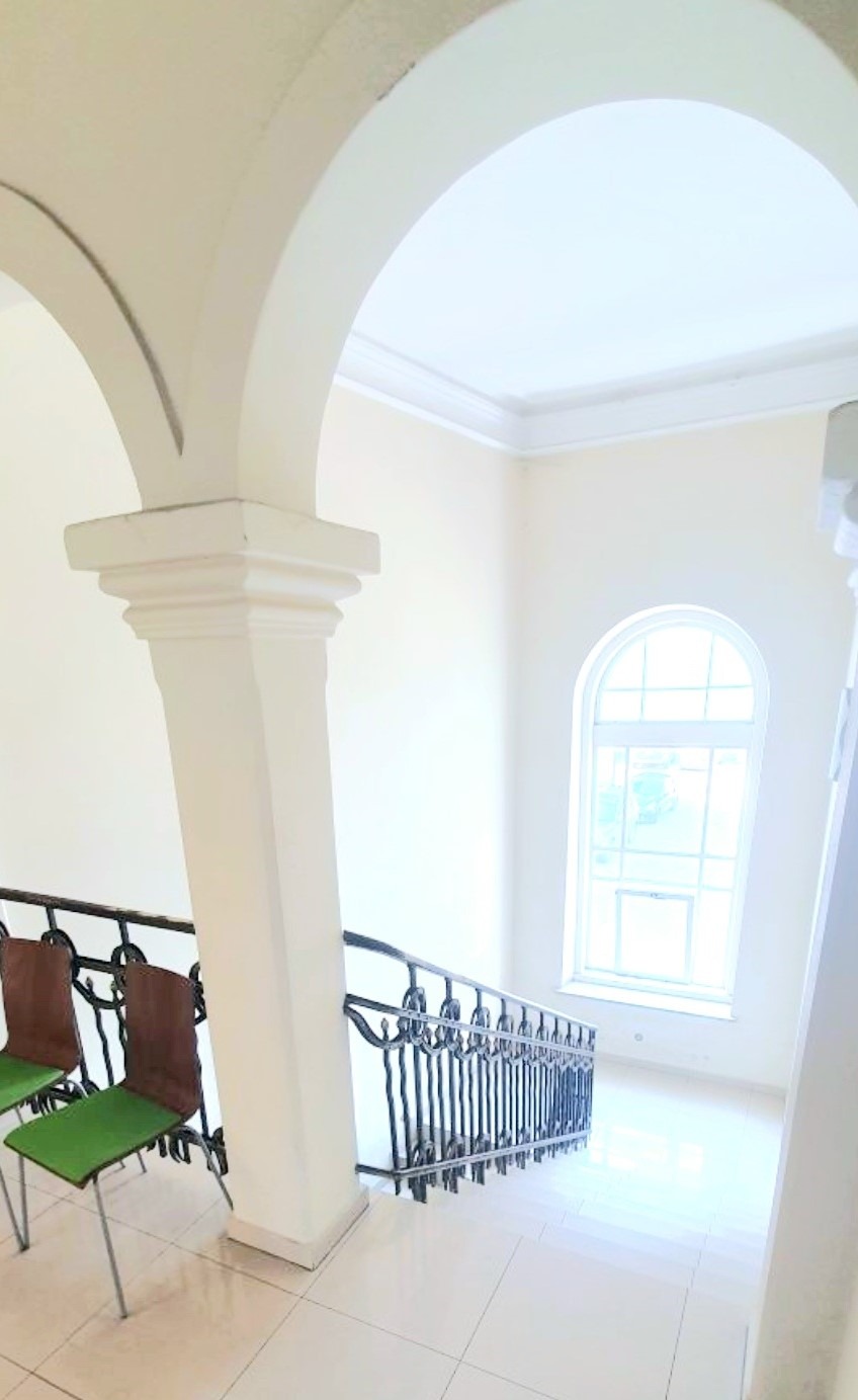 Jasne wnętrze z łukami i schodami. Balustrada z kutego żelaza, okno łukowe. Dwa krzesła z zielonymi siedziskami. Neutralna kolorystyka ścian i podłóg.