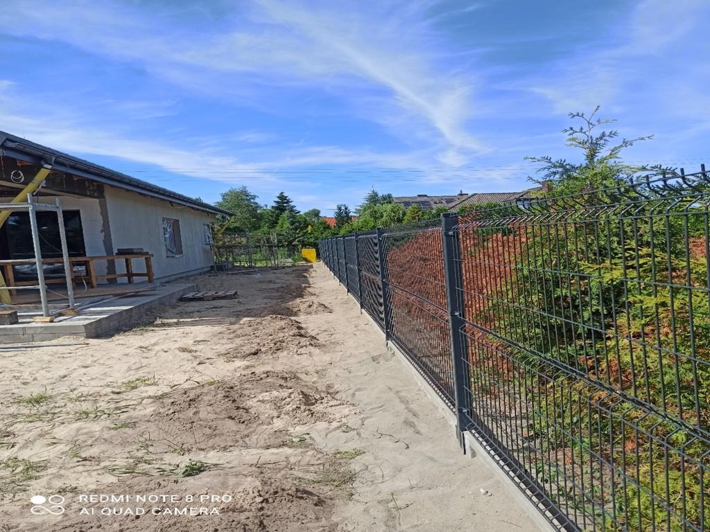 Nowoczesne ogrodzenie panelowe w kolorze antracytowym, zamontowane przy posesji z budynkiem w trakcie wykończenia. Widoczne słupki i podmurówka.