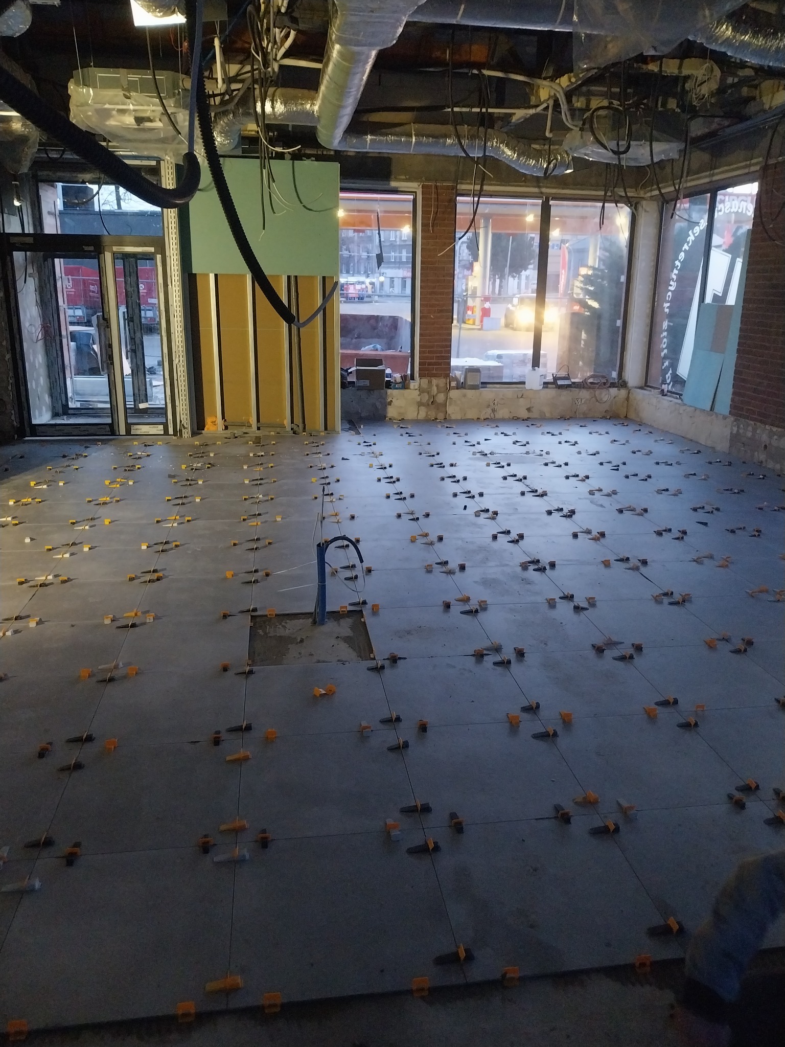 Szare płytki podłogowe w trakcie układania, z pomarańczowymi krzyżykami dystansowymi, w surowym wnętrzu z widocznymi instalacjami pod sufitem i dużymi oknami.