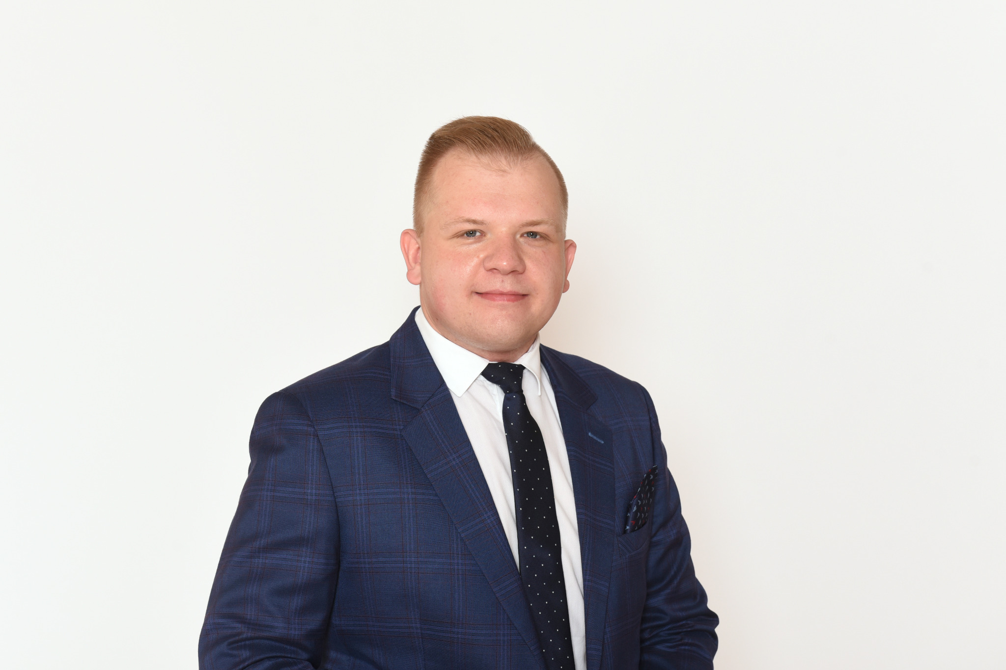 Portret mężczyzny w niebieskiej marynarce w kratę, białej koszuli i krawacie w kropki na jasnym tle. W kieszonce marynarki widoczna poszetka.