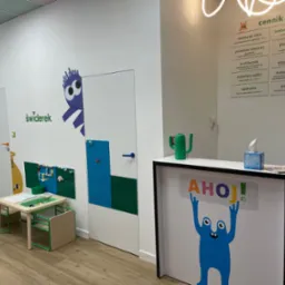Kolorowe wnętrze gabinetu stomatologicznego dla dzieci: recepcja z grafiką, cennik usług, stolik z zabawkami i dekoracje ścian z motywami bajkowymi.
