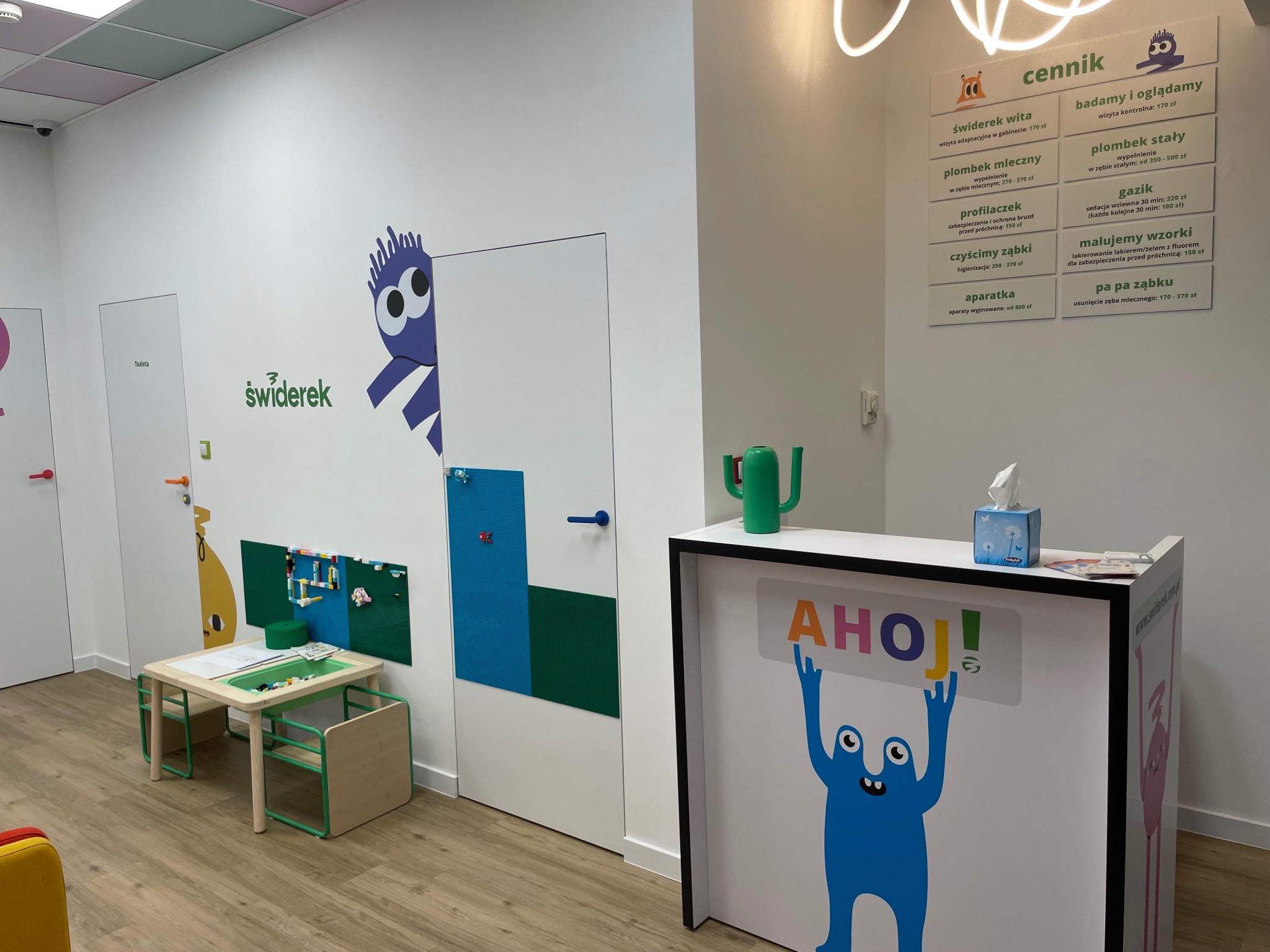 Kolorowe wnętrze gabinetu stomatologicznego dla dzieci: recepcja z grafiką, cennik usług, stolik z zabawkami i dekoracje ścian z motywami bajkowymi.