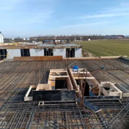 Zbrojenie stropu budynku w trakcie budowy, widok z góry. Stalowe pręty tworzą siatkę z drewnianymi szalunkami. W tle widać budynki i pola.