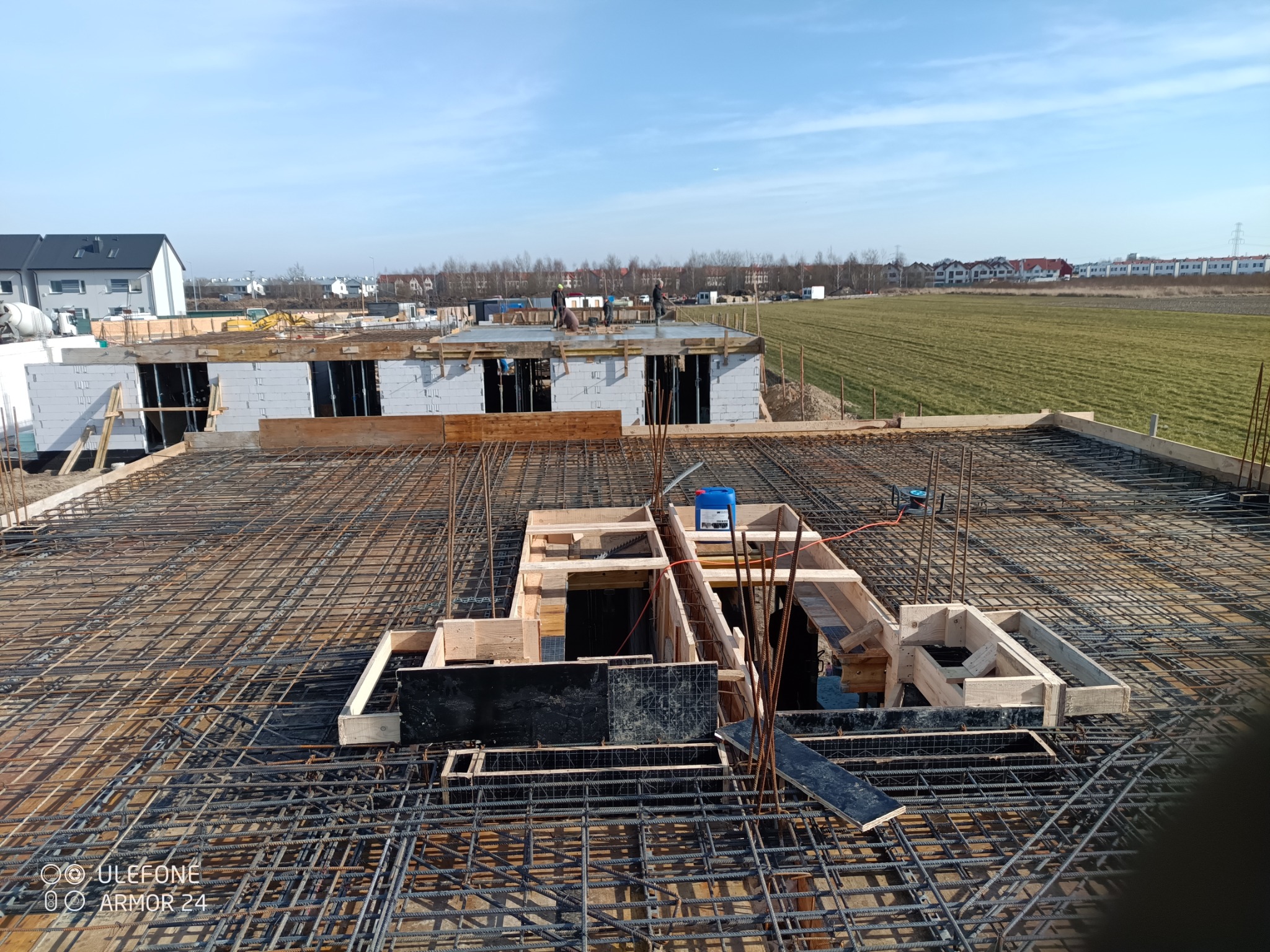 Zbrojenie stropu budynku w trakcie budowy, widok z góry. Stalowe pręty tworzą siatkę z drewnianymi szalunkami. W tle widać budynki i pola.